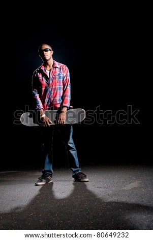 African American skateboarder wearing sunglasses holding his skateboard under dramatic lighting with dramatic shadows.