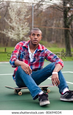 A young man hanging out in the tennis courts sitting on his skateboard.