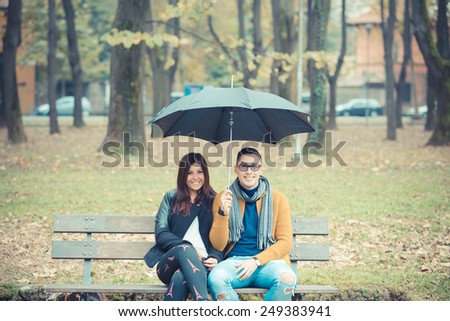 young couple in the park during autumn season outdoor - lovers valentine under umbrella sitting in a bench
