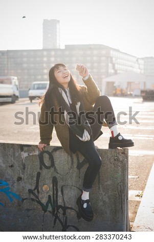 young beautiful asian hipster woman in the city - living town during sunset in backlit - sitting on a wall with smartphone