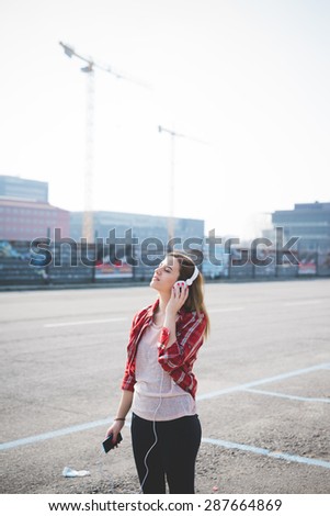 young beautiful blonde hipster woman in the city listening to music with headphones and smartphone