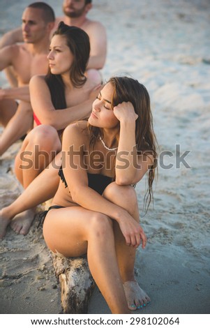 Group of young multiethnic friends seated on the foreshore in swimsuit looking at the horizon at the beach at sunset - future, relax, friendship concept