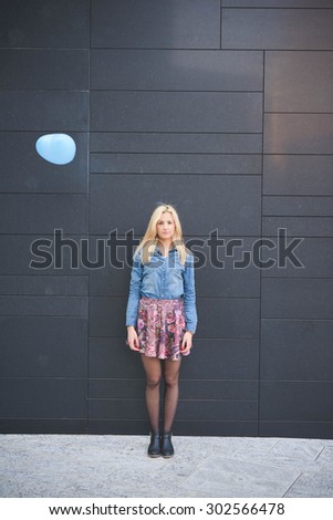 Young beautiful blonde girl posing leaning on a wall with a hearted balloon flying outdoor in the city looking in camera wearing a jeans shirt and a floral skirt