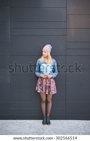 Young beautiful blonde girl posing leaning on a wall with a hearted balloon outdoor in the city overlooking on her right wearing a jeans shirt, a hat, and a floral skirt