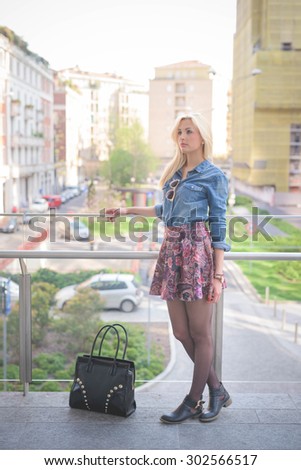 Young beautiful blonde caucasian girl posing outdoor in the city leaning on a windowsill wearing a jeans shirt, a bag and a floral skirt overlooking on her right