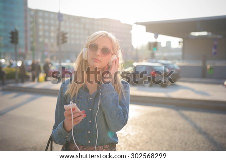 Half length of a young beautiful caucasian blonde girl listening to music in the city with headphones  looking in camera,- relax, youth, emancipation concept