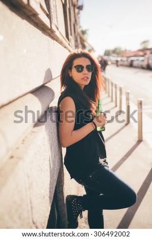 Knee figure of a young beautiful reddish brown hair caucasian woman leaning against a wall drinking a beer - carefreeness, freshness, youth concept - dressed with blue jeans and black shirt