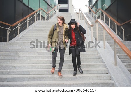 Young couple of caucasian woman and man, with skate and moustache, walking down the stairs hugging and smiling - youth, carefree, freshness concept
