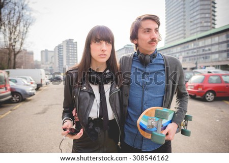 Half length of a couple of young caucasian woman and man with a skate and moustache posing trough the streets listening to music by technological device - carefreeness, friendship, love concept