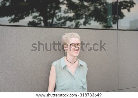 Half length of a young handsome caucasian blonde designer posing leaning against a wall, overlooking left, smiling - wearing a speckled pair of glasses and an azure dress - carefreeness, youth concept