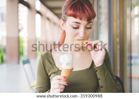 half length of a young handsome caucasian redhead woman eating an ice cream, biting a wafer, looking downward - childhood, freshness concept - wearing a green shirt