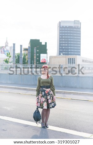 young handsome caucasian redhead woman posing in the city, looking in camera, bag handheld, smiling - carefreeness, youth concept - wearing green shirt, floral skirt and bag