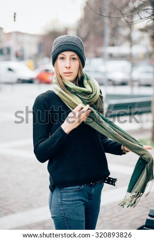 Knee figure of young beautiful hipster sporty blonde woman posing in the city, wearing a scarf, looking in camera, pensive - wearing black sweater, blue jeans and a hat