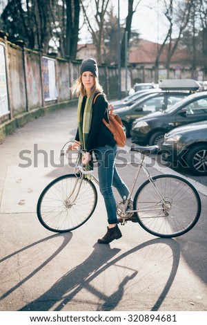 young beautiful hipster sporty blonde woman in town riding her bike, looking in camera, serious - alternative methods of transportation concept - wearing green scarf, blue jeans, black sweater and hat