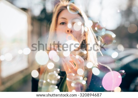 Half length profile of young beautiful hipster sporty blonde woman in town playing with bubble soap, looking in camera, blowing - childhood, happiness, carefreeness concept - backlight