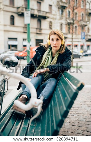 young beautiful hipster sporty blonde woman lying down on a bench, with bike and headphones, listening music, overlooking right - relaxing, music concept