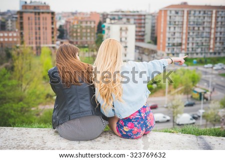 Two young blonde and brunette girls chatting sitting on a small wall with city on background, view from back, one indicating something on left - friendship, carefreeness concept