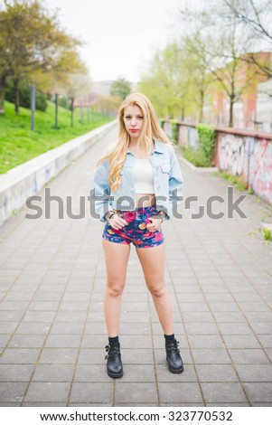 Young handsome blonde caucasian fashion girl posing on a park in the city suburbs, hands in pocket, looking in camera - carefreeness, youth concept - wearing jeans jacket and floral shorts