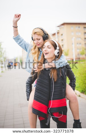 Knee figure of two young blonde and brunette girls listening to music in the city having fun, smiling - music, relaxing, technology, friendship concept - blonde piggyback on back on her friends