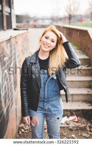 Knee figure of young beautiful blonde straight hair woman in the city, touching her hair, smiling, looking in camera - happiness, carefreeness concept - wearing jeans overalls, black leather jacket