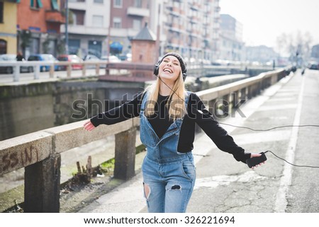 Knee figure of young beautiful blonde straight hair woman in the city with headphones listening to music, arms wide open, feeling free, laughing - freedom, happiness concept