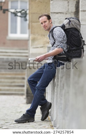 Side view portrait of male hiker holding book while sitting on retaining wall in city