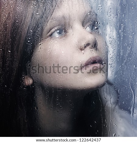 Art portrait of a beautiful young sadly girl standing at the window watching the rain