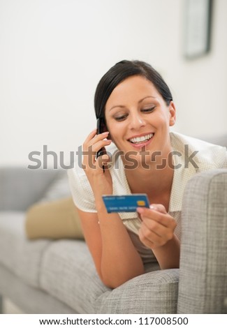 Happy young woman on sofa holding credit card and speaking mobile phone