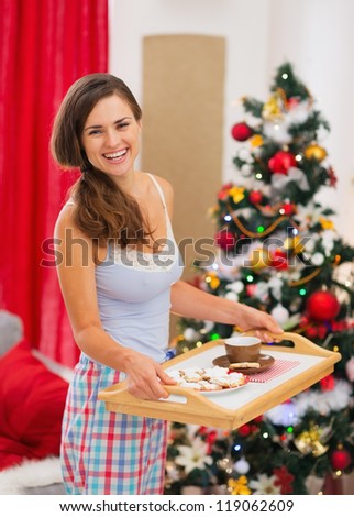 Happy woman in pajamas holding bed table with snacks in front of Christmas tree