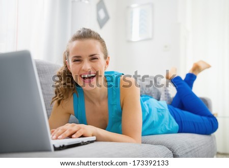 Smiling young woman laying on sofa and using laptop