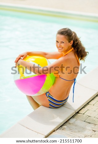 Portrait of smiling young woman with ball sitting near swimming pool