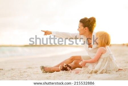 Baby girl and mother pointing on copy space while sitting on the beach in the evening