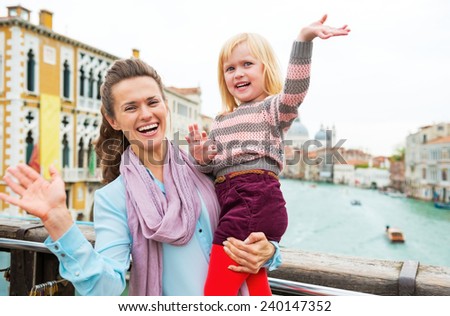 Happy mother and baby girl waving while standing on bridge with grand canal view in venice, italy