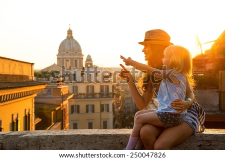 Mother and baby girl sitting on street overlooking rooftops of rome on sunset and pointing