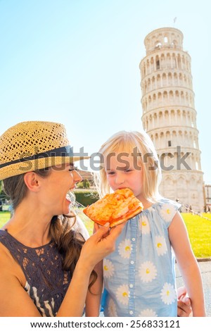 Happy mother and baby girl eating pizza in front of leaning tower of pisa, tuscany, italy
