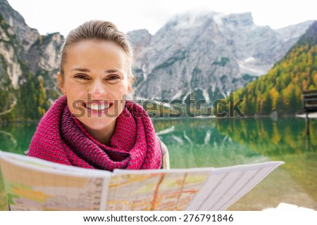 Portrait of a smiling brunette hiker holding a map. In the background, autumn colours and the Dolomite mountains are reflecting in the still water of Lake Bries. Autumn colours and leaves.