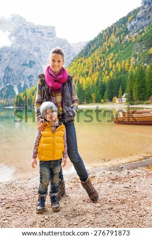 A mother and daughter standing on the shore of Lake Bries wearing outdoor gear. In the background autumn colours, green trees, and wooden boats are reflecting in the water.