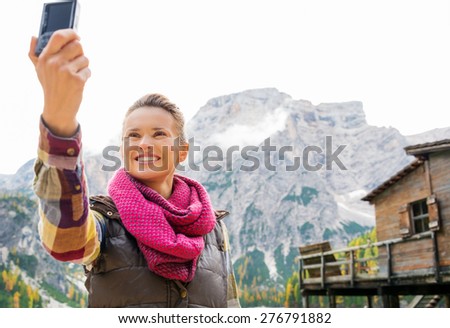 A smiling brunette wearing outdoor gear is taking a self-portrait. The Italian Dolomites are in the background, as is a rustic wooden pier and house.