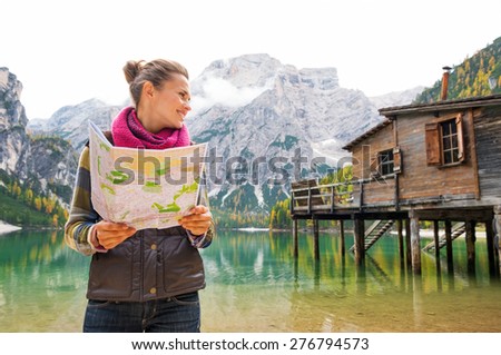 An smiling brunette wearing outdoor gear is looking sideways, holding an open map in her hands. Behind her, the Italian Dolomites are reflected in the still water.