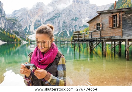 A smiling brunette wearing outdoor gear looks at the photo she has taken on her digital camera. In the background, the Italian Dolomites are above the lake.