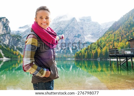 A smiling brunette is pointing into the distance at the scenery while smiling. In the background, autumn colours, the Dolomite mountains and wooden pier are reflected in the water.