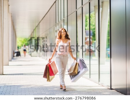 A brown-haired woman wearing muted, gentle colours holds five colourful, patterned shopping bags walks towards the doors of an exclusive shopping mall.