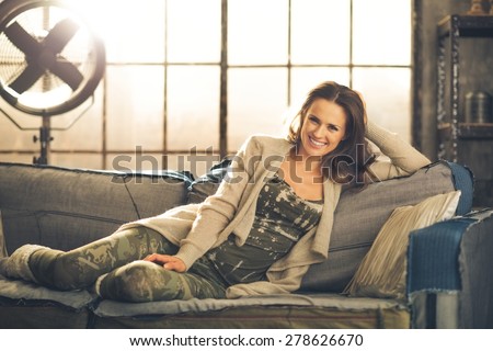 A brunette woman is smiling, relaxing on a sofa, feet curled under her, wearing comfortable clothing, leggings, and a cardigan. Industrial chic ambiance and cozy atmosphere.
