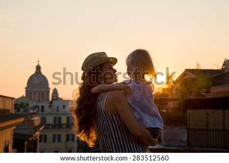 At sunset, with views over Rome, a brunette mother wearing a hat is holding her daughter - seen from behind - in her arms, and talking to her. In the distance, St. Peter\'s Basilica.