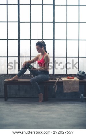 A healthy, strong woman is sitting casually on a bench, holding her device. Looking down at it, she is selecting the kind of music she would like to listen to for her workout.