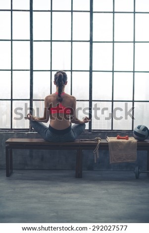 Sitting on a bench by a big window in a city loft gym, a woman is in lotus position, meditating.