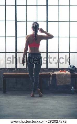 A fit, strong, muscular woman is standing, legs crossed, while looking out the window. In one hand, she is holding her device, listening to music. With the other hand, she is adjusting her earbud.