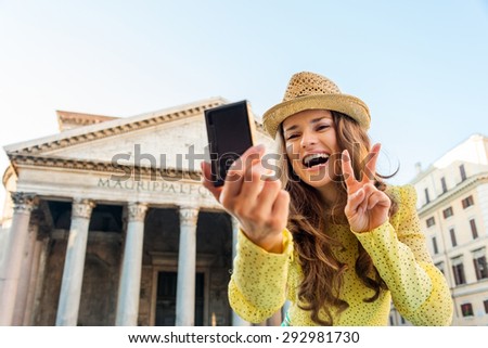 A woman tourist smiles as she is taking a selfie, doing the victory sign. In the background, the Pantheon in Rome on a summer\'s day.