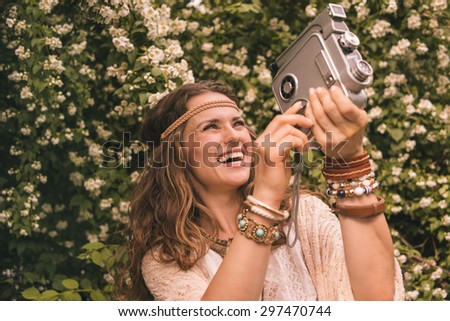 Longhaired hippy-looking young lady in knitted shawl and white blouse standing among flowers with retro camera