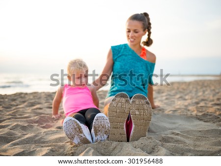 A fit, healthy young mother is sitting in the sand on the beach at sunset. They are comparing shoe sizes.
